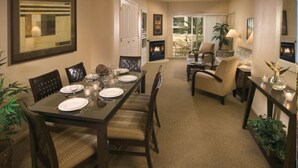 A dining area with a long table set for four, surrounded by chairs. There is a fireplace in the background, and the room has a carpeted floor.
