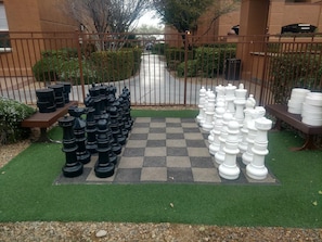 Un jeu d’échecs installé sur une pelouse artificielle, avec des pièces noires d’un côté et blanches de l’autre, de part et d’autre d’une clôture métallique.