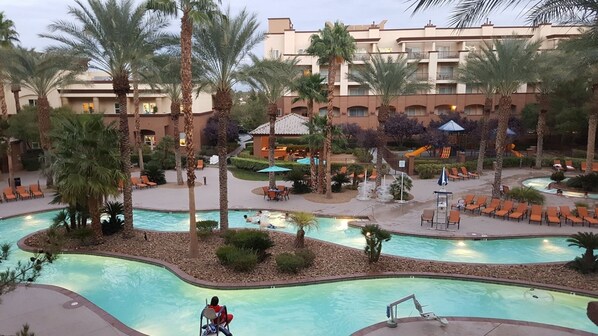 Ein Poolbereich eines Resorts mit mehreren Swimmingpools, Palmen, Liegestühlen und einem Gebäude im Hintergrund.