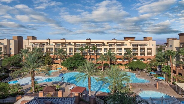 Un hotel con varios balcones, una piscina grande en el centro y palmeras que lo rodean.