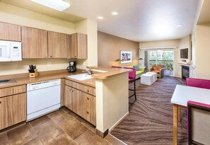 A modern kitchen with a dishwasher, a coffee maker, and a microwave. There is a dining area with a sofa and a TV.