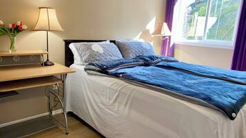 A neatly made bed with a blue blanket and pillows, a bedside table with a lamp, and a window with purple curtains.