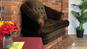 Una habitación acogedora con pared de ladrillo, un sillón mullido, una pequeña mesa con mantel rojo, una planta en maceta y fotografías enmarcadas en la pared.