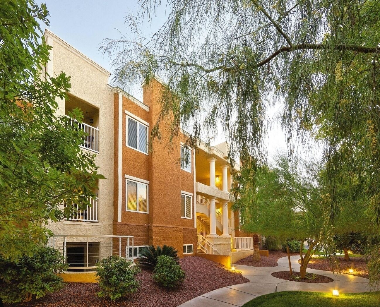 A multi-story building with a brick facade, balconies, and a landscaped courtyard with trees and shrubs.