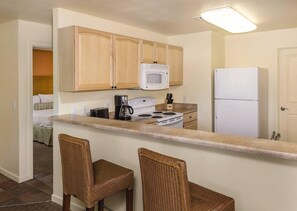 Una cocina compacta con armarios de madera, un microondas, una estufa, un refrigerador y dos taburetes de mimbre.