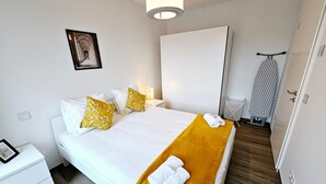Ein ordentlich bezogenes Bett mit weißen Laken und einer senfgelben Tagesdecke, ein Nachttisch mit Lampe sowie ein an der Wand hängendes Bild.
