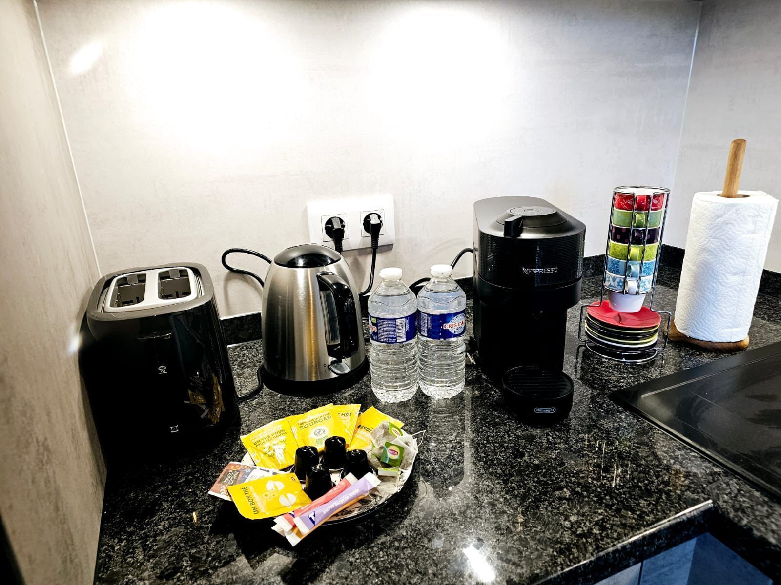A kitchen counter with a toaster, kettle, coffee maker, water bottles, and a plate with condiments.