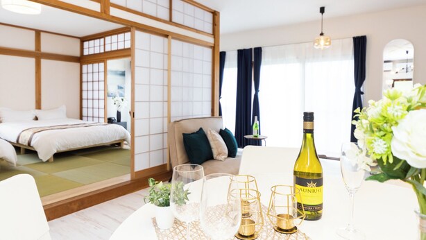 A modern hotel room with a traditional Japanese sliding door, a bed, a sofa, a dining table with a bottle of wine, glasses, and a vase of flowers. - Awaji Large Log House in Goshiki 2 (Sumoto)