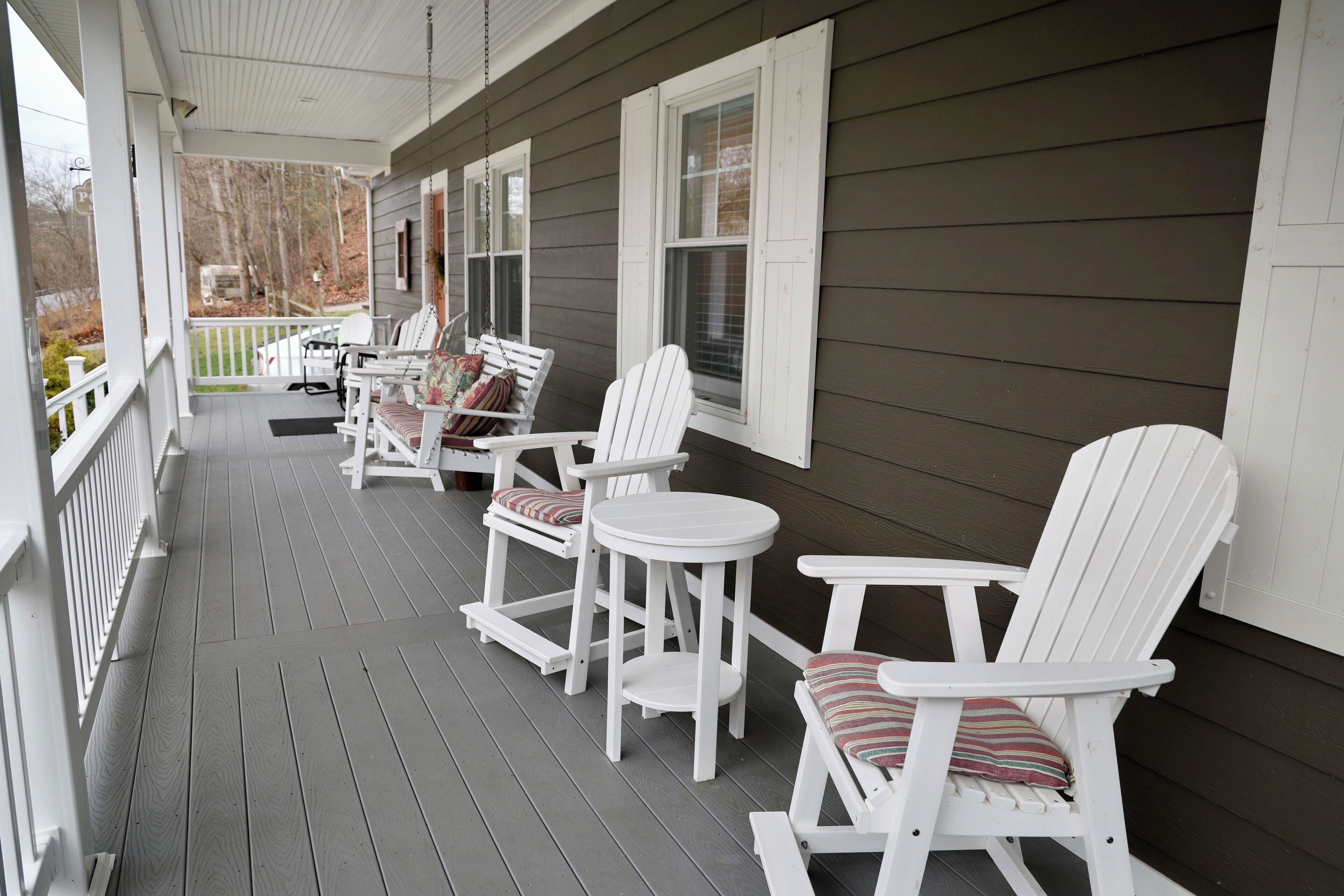 A porch with white Adirondack chairs and a small round table, a white railing, and a view of the yard.