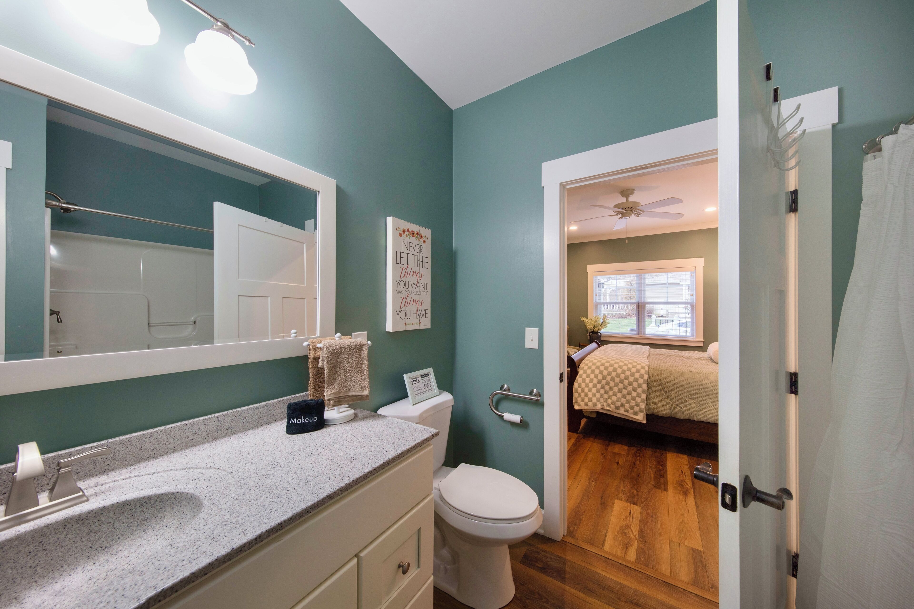 Een badkamer met een grote spiegel, een wastafel en een toilet. Er is een handdoekhouder en een bordje aan de muur. De badkamer heeft muurpanelen in teal en houten vloeren.