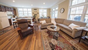 A spacious living room with a brown leather sofa, a beige couch, and a coffee table. A kitchen with wooden cabinets and a dining area with a table and chairs is visible in the background.