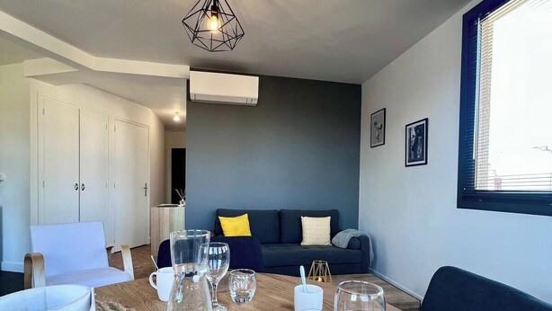 A modern living room with a round wooden table, a dark blue sofa, and a window with blinds. - Casa #1407 - T2 - Climatisé avec Balcon - Centre-ville (Brive-la-Gaillarde)