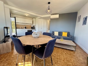 Un espacio moderno para vivir con una mesa redonda de madera, sillas azul oscuro, un sofá gris con almohadas amarillas y una cocina al fondo.