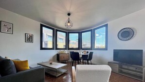 A modern living room with a sofa, a coffee table, a dining area with chairs, a TV, and large windows with blinds. - Casa #1407 - T2 - Climatisé avec Balcon - Centre-ville (Brive-la-Gaillarde)