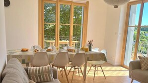 Un salon moderne avec un canapé, une table à manger et des chaises. De grandes fenêtres offrent une vue sur la verdure environnante.
