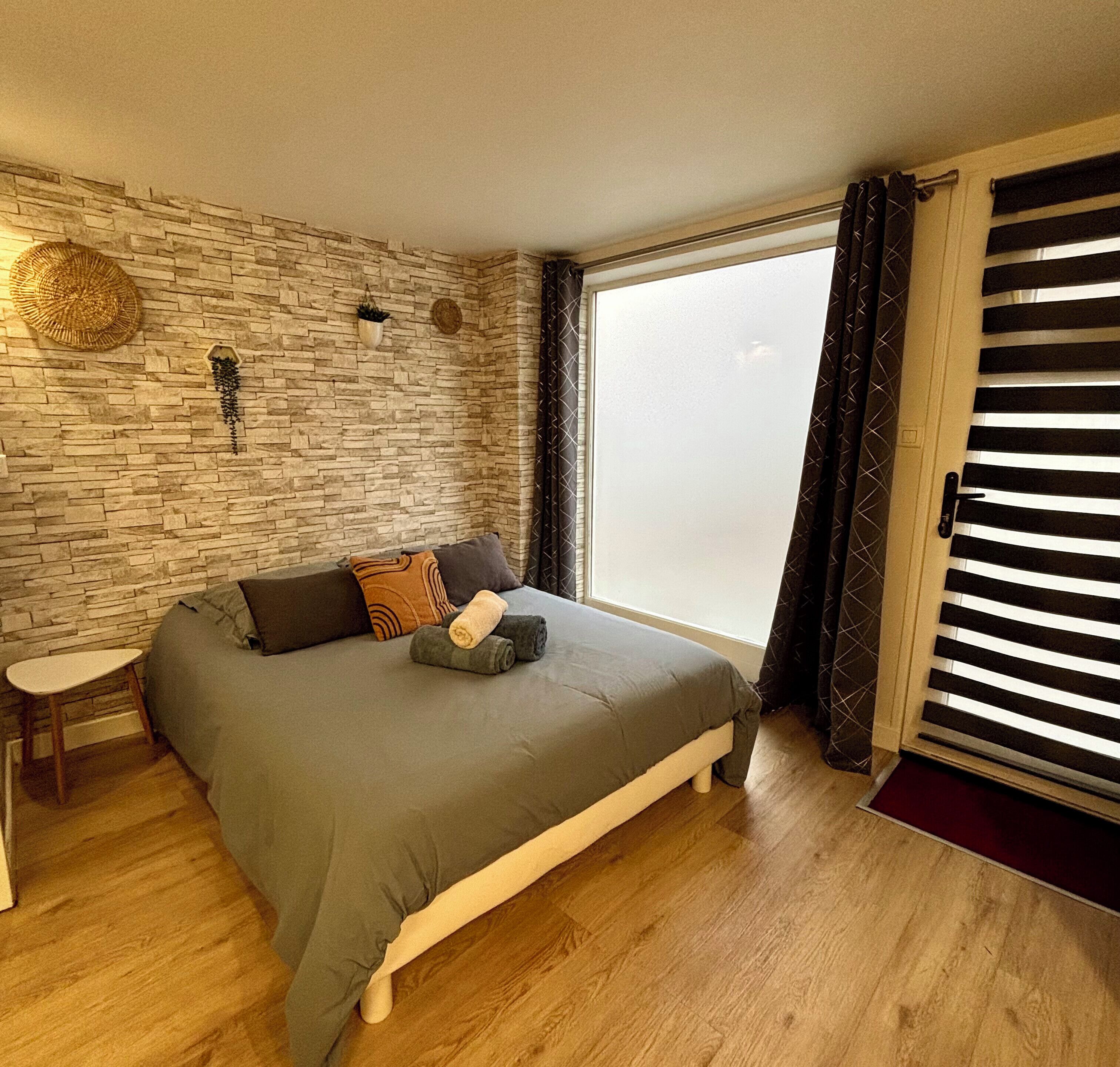 A bedroom with a stone wall, a bed with grey bedding, a small bedside table, and a window with curtains.