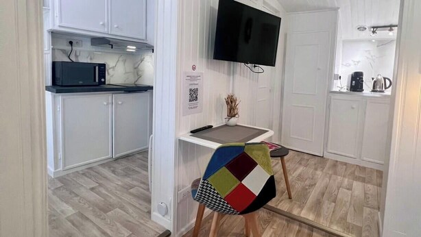 Una cocina compacta con muebles blancos, microondas y cafetera. Un televisor está montado en la pared sobre una pequeña mesa.