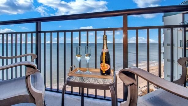 En balkon med et bord, der holder en champagneflaske og to glas, med udsigt over en strand og et hav.