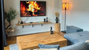 Ein modernes Wohnzimmer mit einem Flachbildfernseher, der ein Kamin zeigt, einem Holztisch, blauen Sofas, einer Stehlampe und einer kleinen Pflanze.