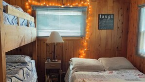 A cozy cabin room with wooden walls, a bunk bed on the left, a single bed on the right, a small table with a lamp, and a sign that says "laugh." - Haven at Loon Lake (Grand Marais)