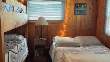A cozy cabin room with wooden walls, a bunk bed on the left, a single bed on the right, a small table with a lamp, and a sign that says "laugh."