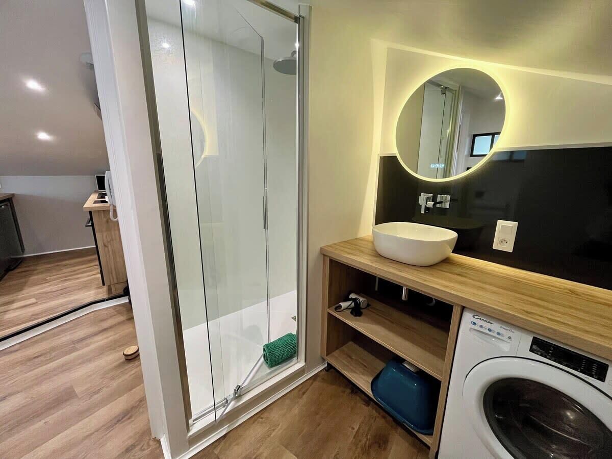 A modern bathroom with a glass shower enclosure, a sink with a round mirror, and a washing machine.
