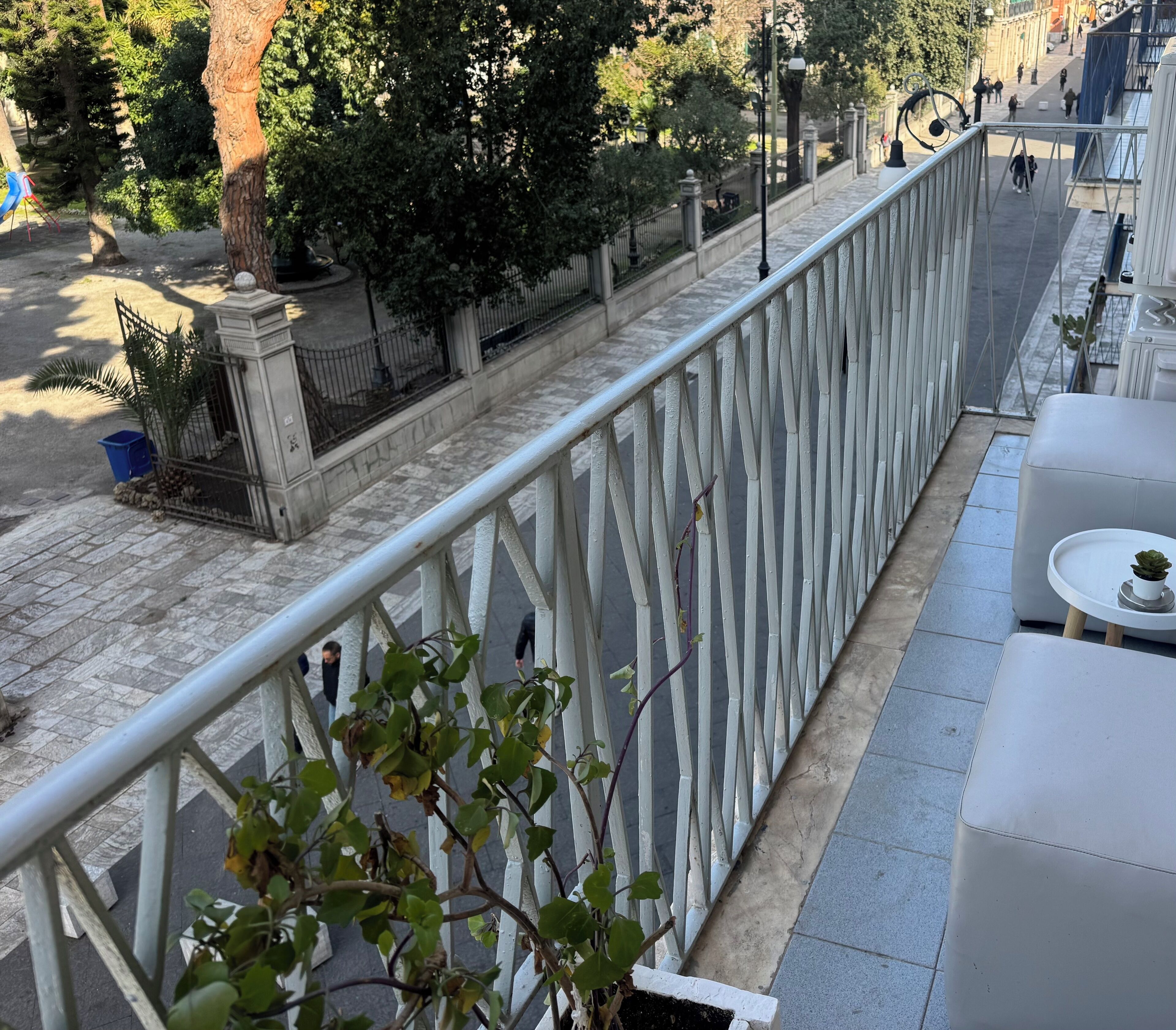 Ein Balkon mit weißer Brüstung, einem Topfpflanzen und einer Sitzecke mit Tisch und Stühlen.
