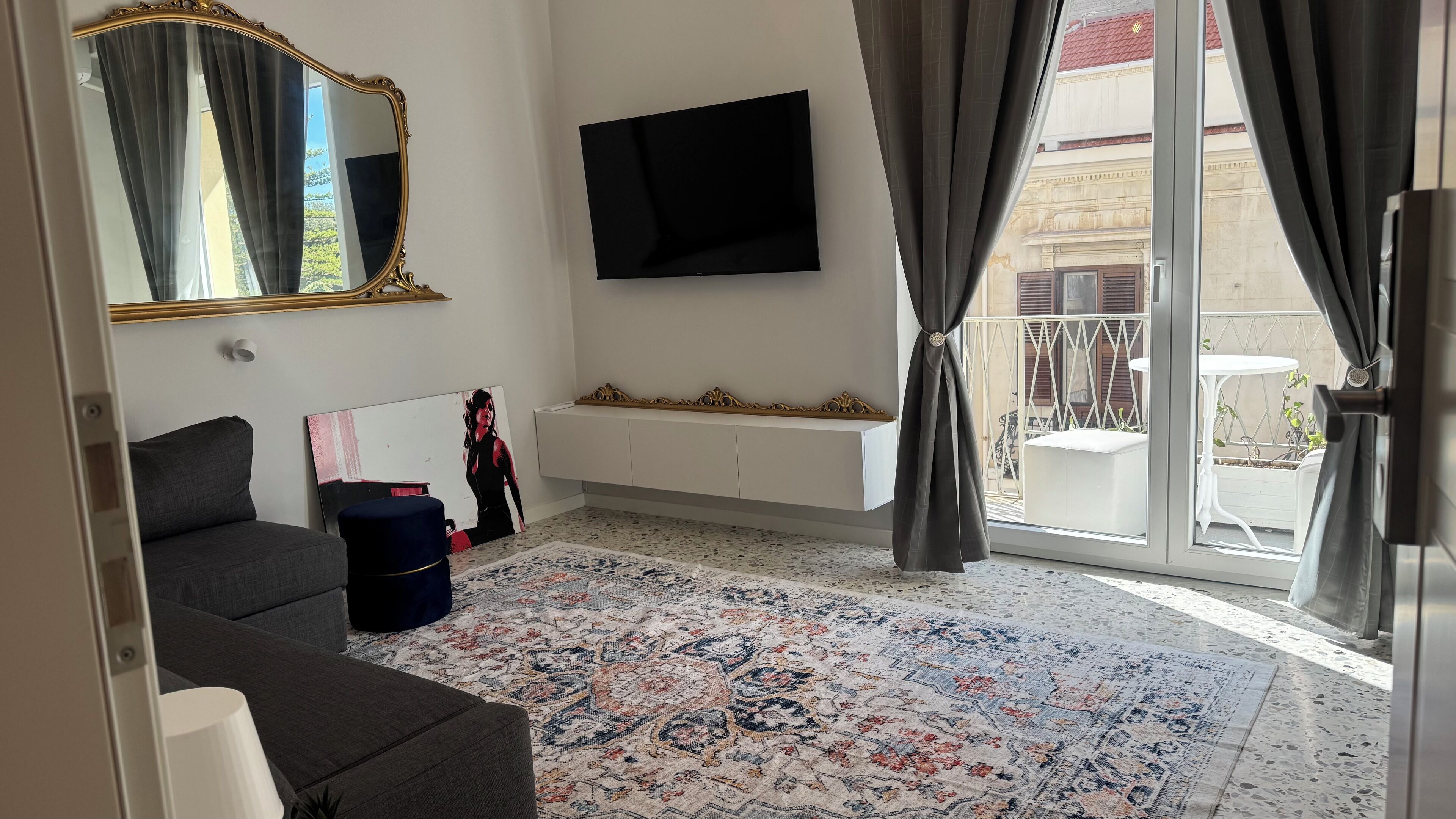 A modern living room with a sofa, a flat-screen TV, a patterned rug, and a balcony with outdoor furniture.