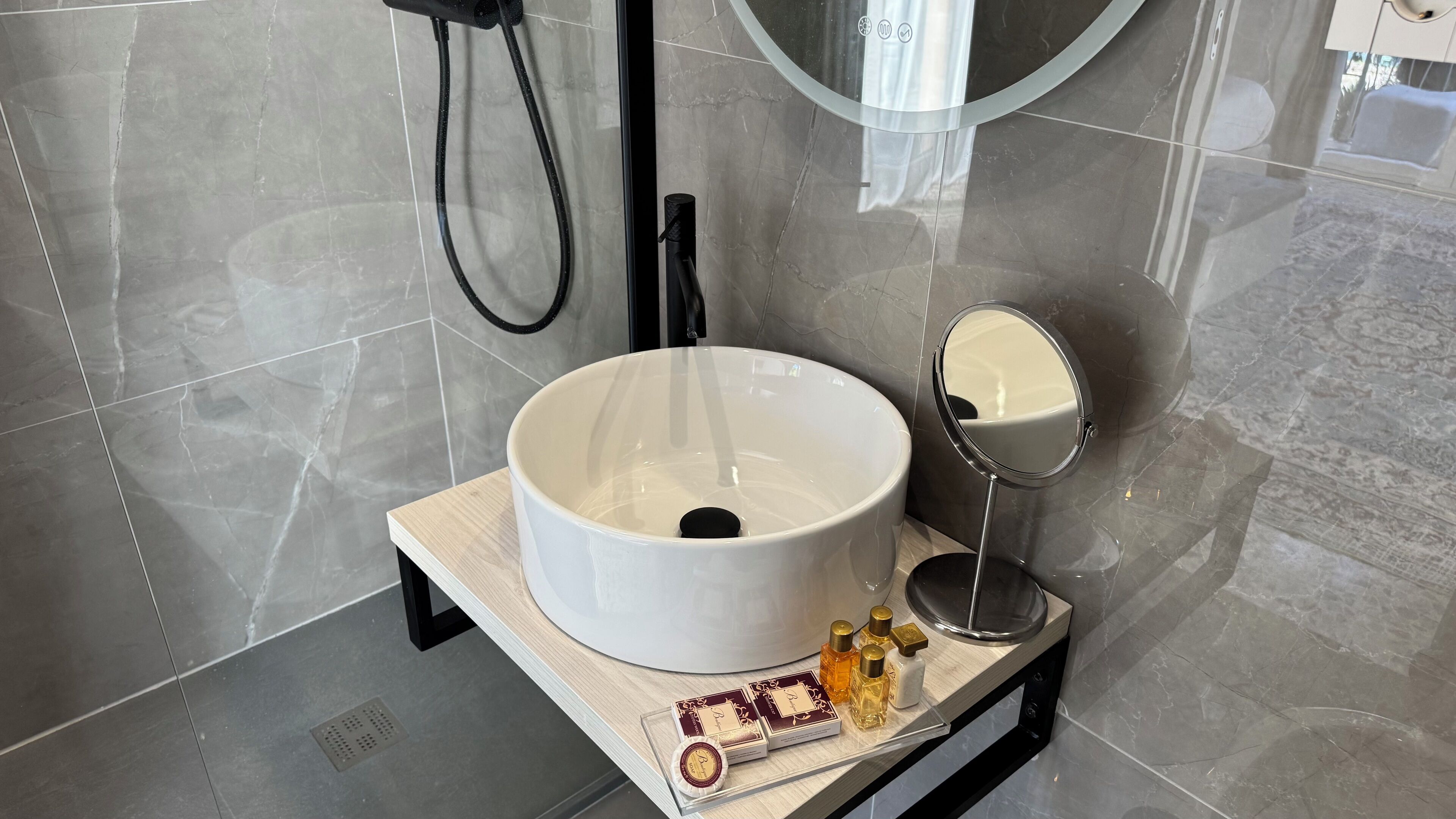 A bathroom with a large white sink, a handheld showerhead, a round mirror, and a small tray with toiletries.