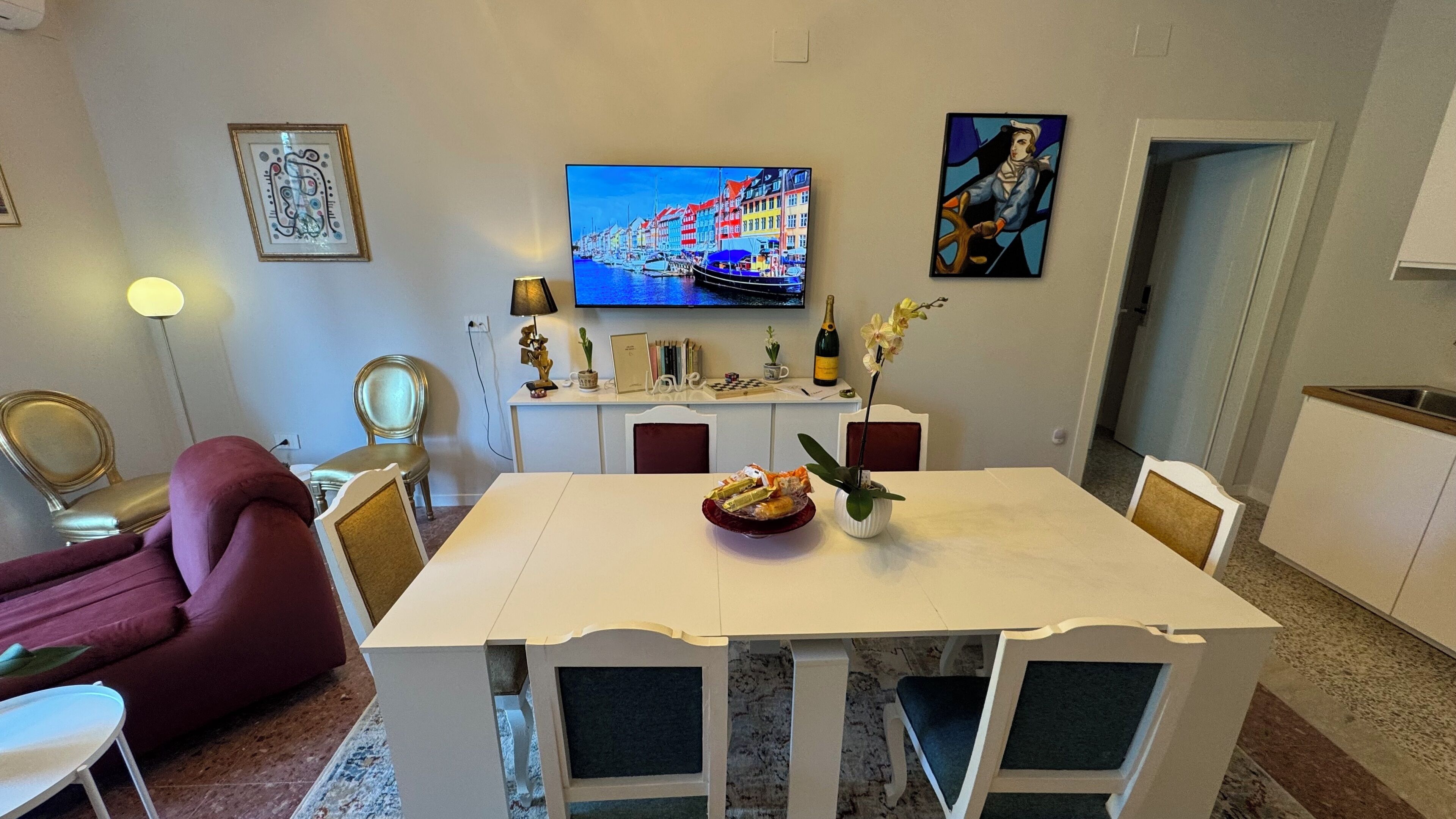 Un coin repas avec une table blanche, des chaises et un vase rempli de fleurs. Un téléviseur fixé au mur diffuse une scène aux couleurs vives.