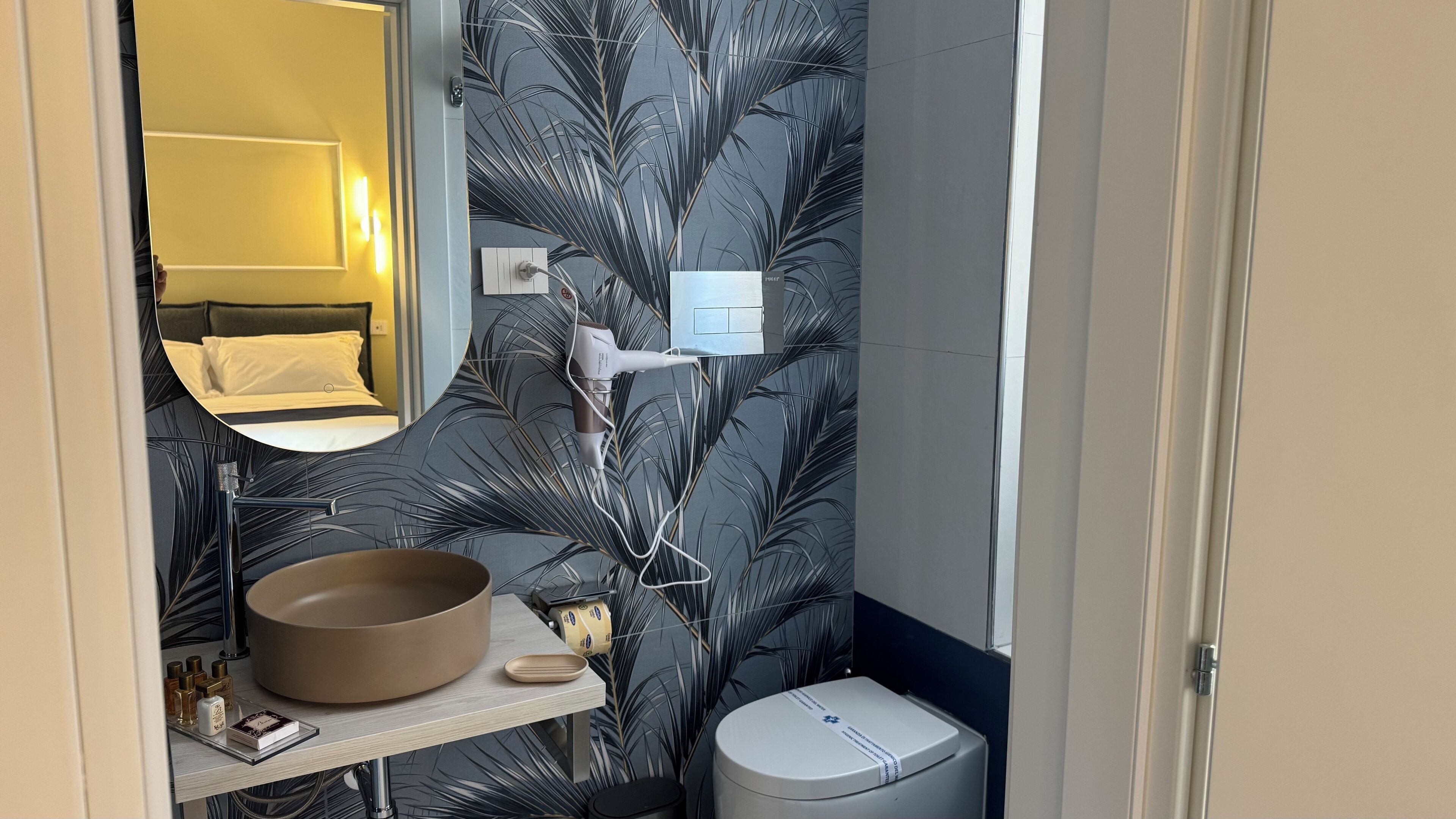 A bathroom with a mirror, sink, and toilet. There is a towel rack above the sink and a towel on the floor. The walls have a leaf pattern.