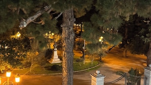Vista nocturna de una zona vallada con un árbol grande, una estatua y una calle iluminada por farolas.