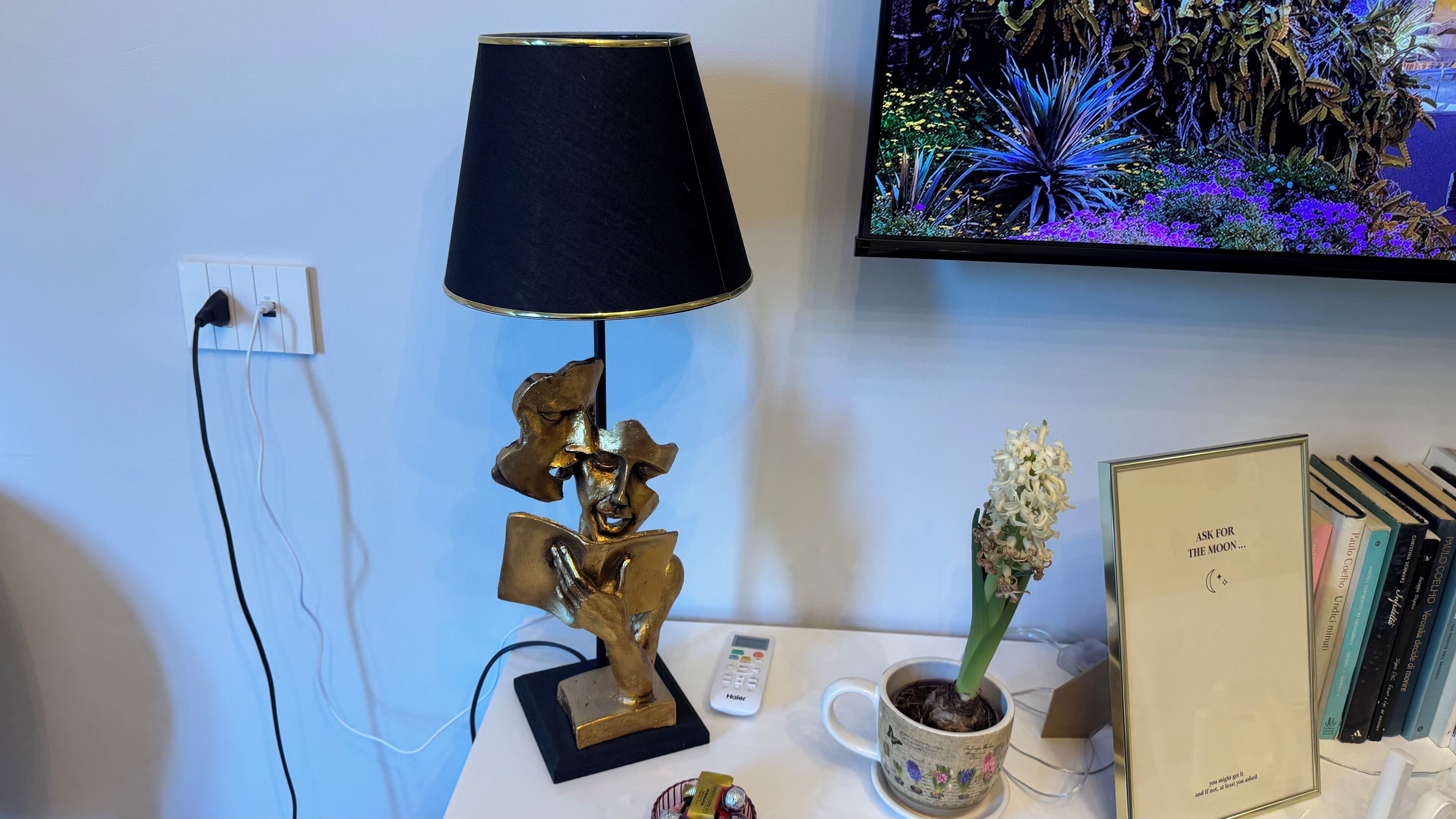 Uma mesa com uma luminária, um vaso de planta, um controle remoto e um livro intitulado "Ask for the Moon...".