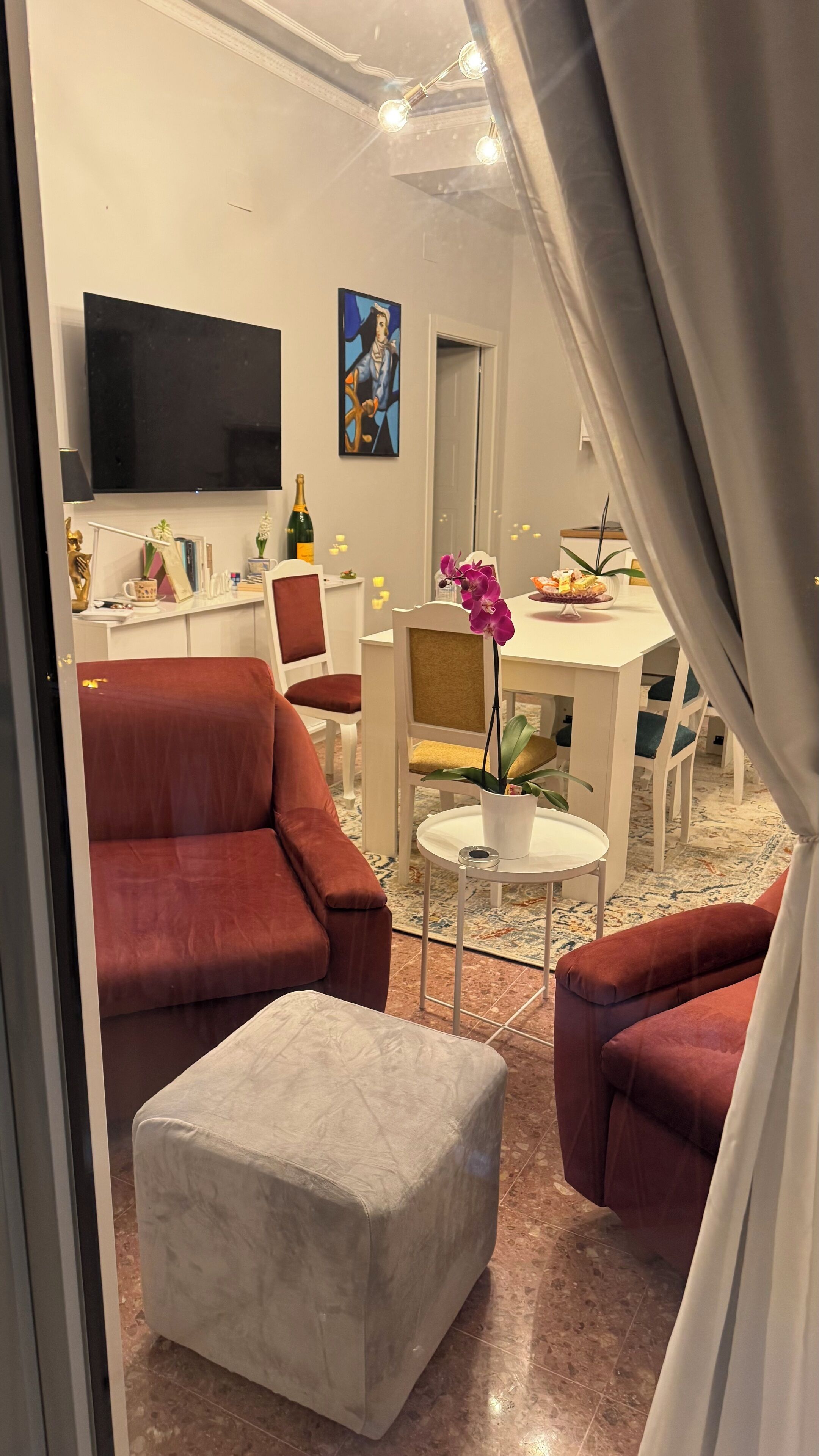 Uma sala de estar com um sofá vermelho, um banquinho branco, uma televisão, um quadro e um espaço de jantar com mesa e cadeiras.