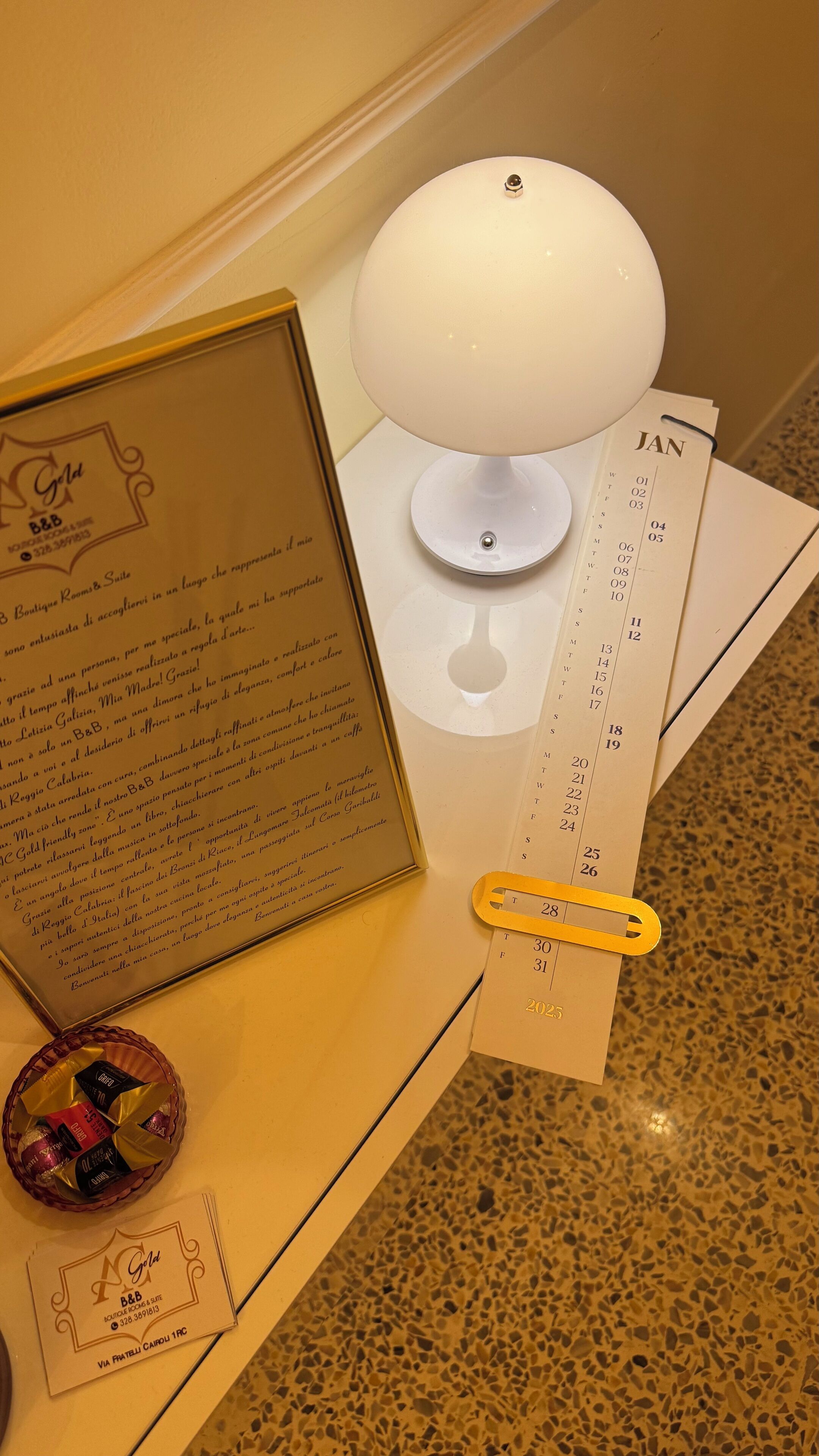 A hotel reception desk with a framed welcome message, a calendar, a lamp, and a small basket of chocolates.