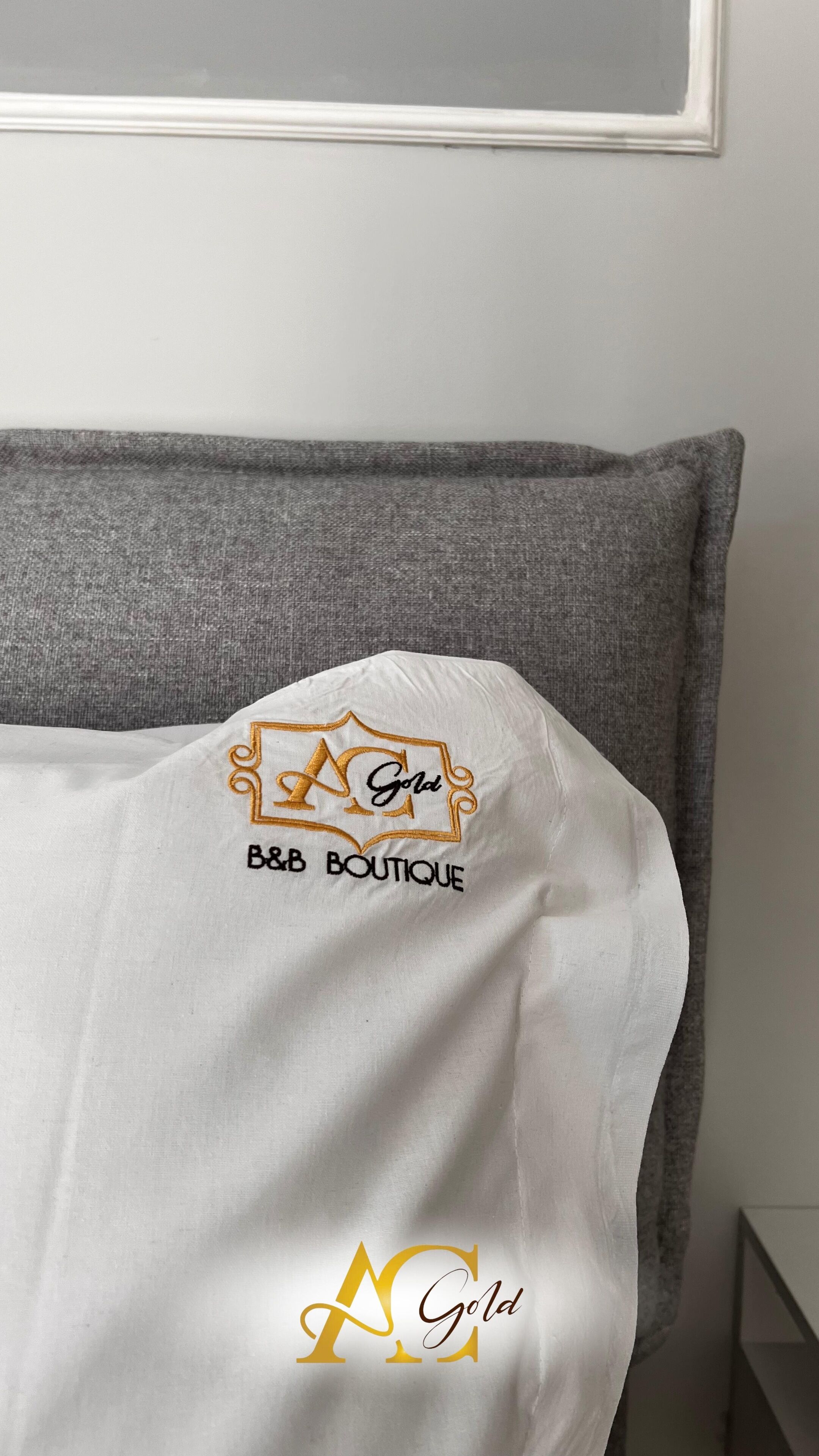 A white bedspread with embroidered text and logo on a grey upholstered headboard.