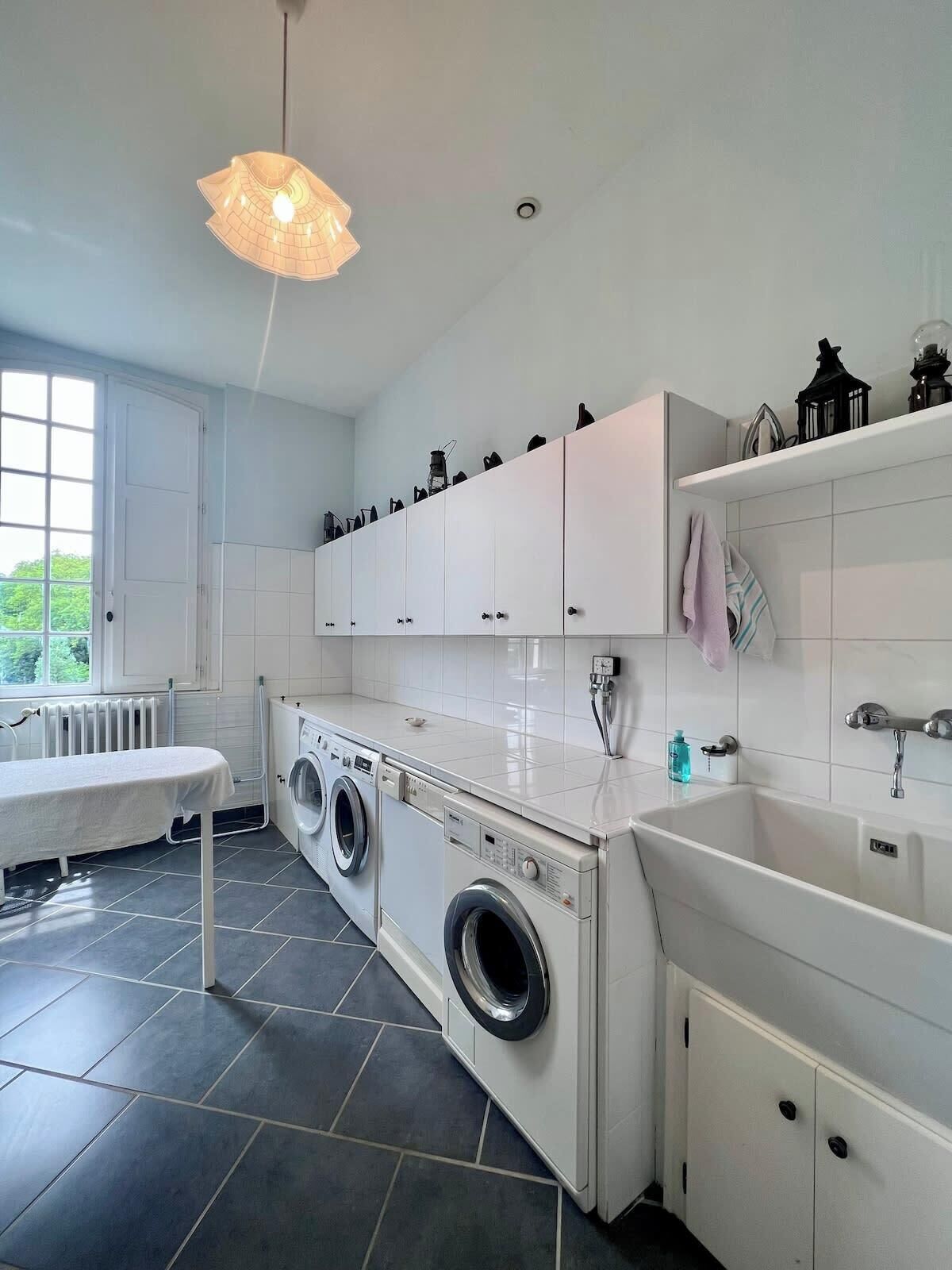 Une buanderie moderne équipée d’un lave-linge, d’un sèche-linge et d’un évier. Une fenêtre laisse entrer la lumière naturelle et un luminaire suspendu est fixé au plafond.