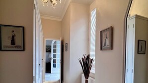 Ein Korridor mit Holzboden, weißen Wänden und einer Chandelier. An den Wänden befinden sich gerahmte Bilder und ein Topfpflanze steht in der Nähe eines Fensters.