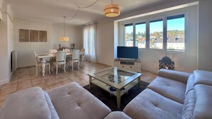 A living room with a glass coffee table, white sofas, a dining area with a table and chairs, a TV on a stand, and a large window with a view of buildings. - Casa Ribot #10 - T4 pour 5 personnes - Proche Centre Ville (Brive-la-Gaillarde)