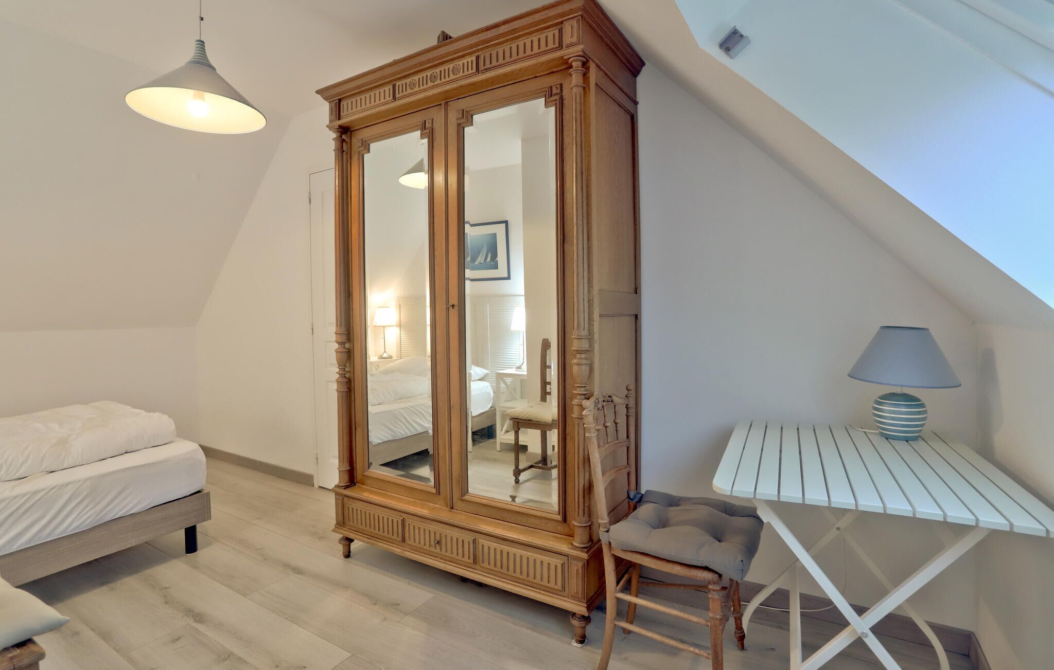 A bedroom with a large mirror, a bed, a desk with a lamp, and a chair.