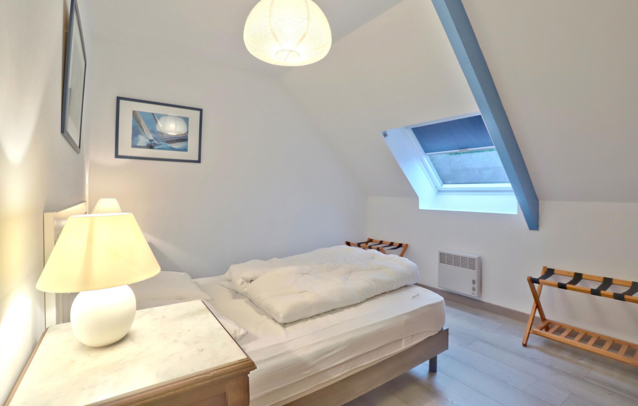 A bedroom with a bed, a nightstand, a lamp, a picture on the wall, a skylight, and a window.
