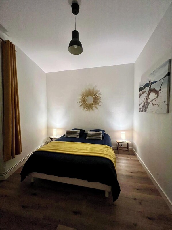 Un dormitorio con una cama, dos mesitas de noche, una lámpara colgante, un espejo adosado a la pared y un cuadro.