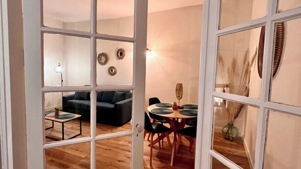 Een kamer met houten vloer, een zwarte bank, een eettafel met stoelen en een witte deur met ruitpanelen.