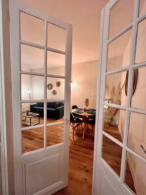 Una habitación con suelo de madera, un sofá negro, una mesa de comedor con sillas y una puerta blanca con paneles de vidrio.