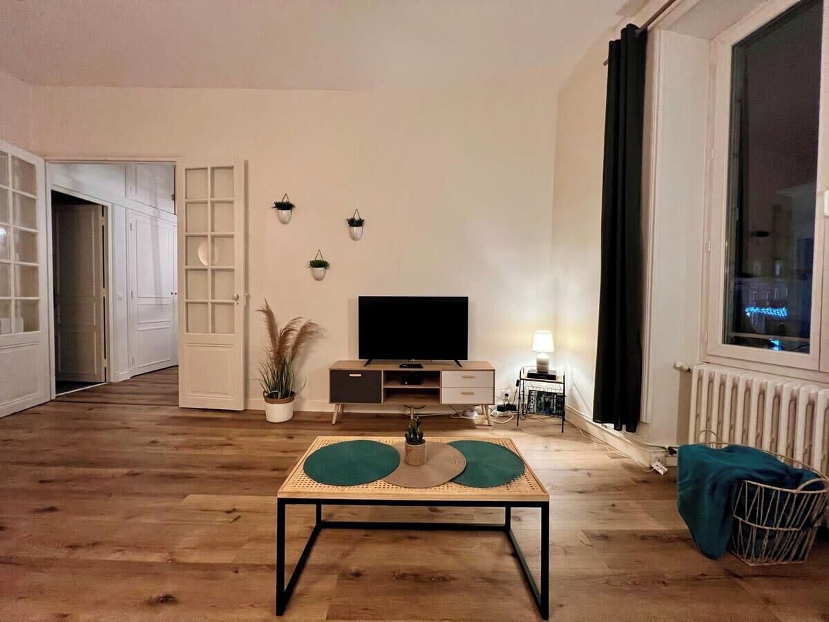 Un salon avec un plancher en bois, un téléviseur posé sur un meuble, une petite table avec une plante et une fenêtre avec des rideaux.