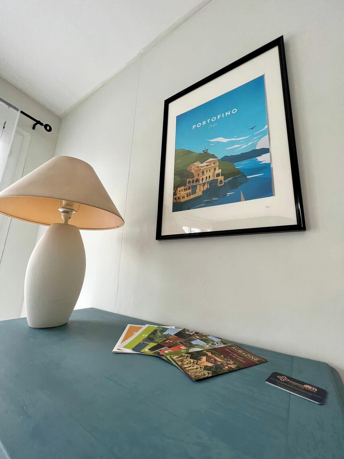 En inramad bild av Portofino hänger på väggen, en lampa står på bordet och en tidning ligger på bordet.