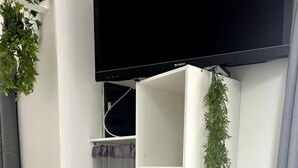 Un espacio habitable compacto con una pared blanca, un televisor de pantalla plana montado sobre un mueble blanco con estanterías, un microondas sobre un gabinete blanco y una planta colgante.