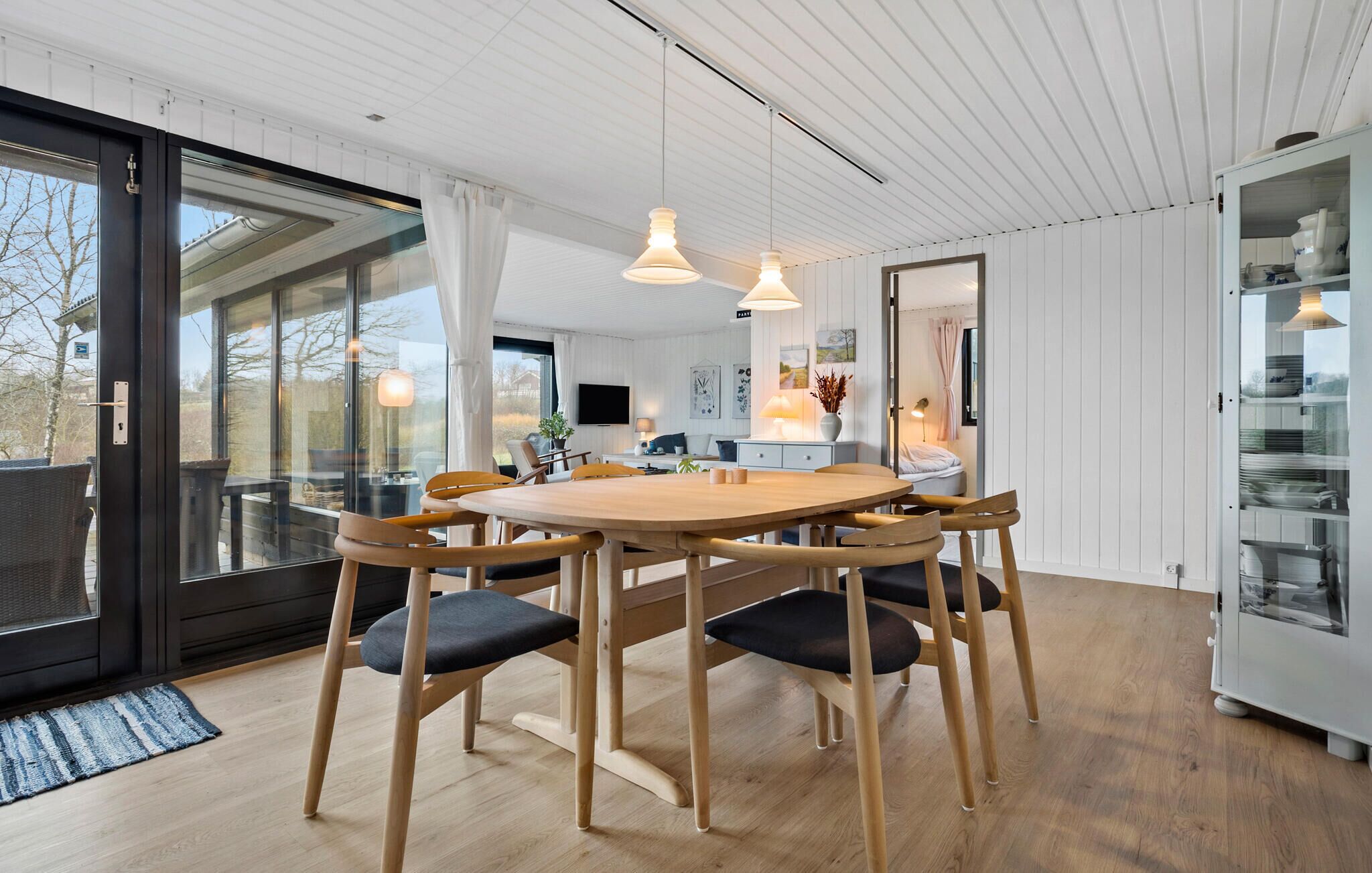 A modern kitchen with a wooden dining table, chairs, pendant lights, and a glass cabinet.