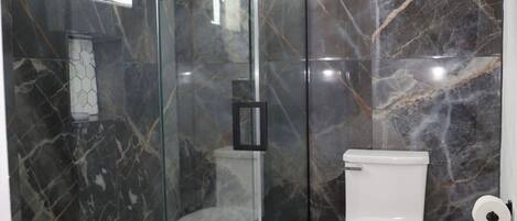 A modern bathroom with a glass-enclosed shower, a toilet, and a trash bin.