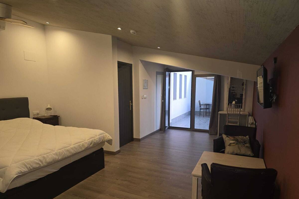 A modern hotel room with a large bed, a desk, and a chair. There is a door leading to a balcony with a view.